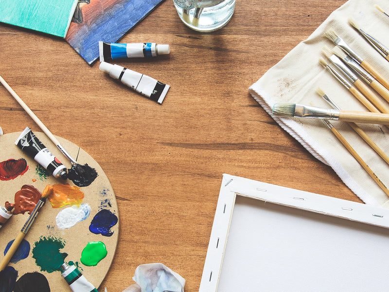 Art by the Lake Schroon art supplies including paint tubes, brushes, canvas, and a palette on a wooden table